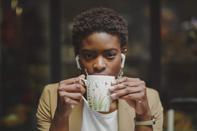 African American woman with earphones looking at camera, holding mug of beverage and sitting on blurred background — Stock Photo