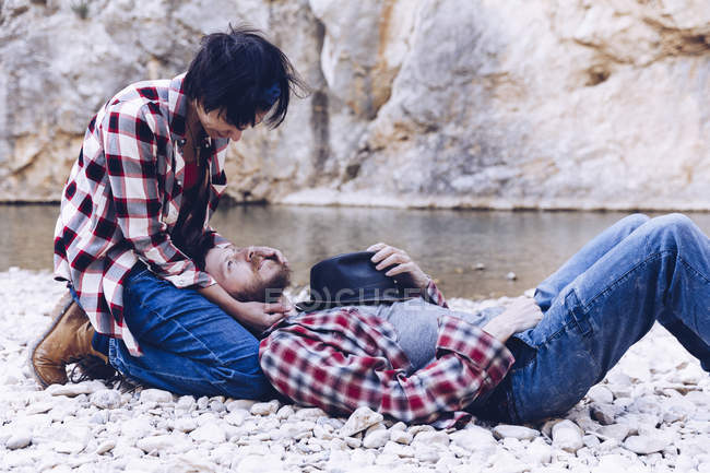 Вид сбоку на молодую пару, лежащую на скалистом берегу горной реки возле скалы — стоковое фото
