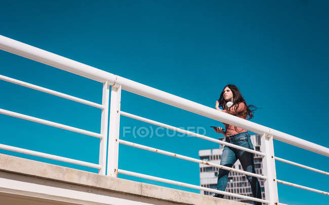 Знизу впевнена молода жінка зі смартфоном і навушниками, що ходять по мосту на фоні блакитного неба — стокове фото