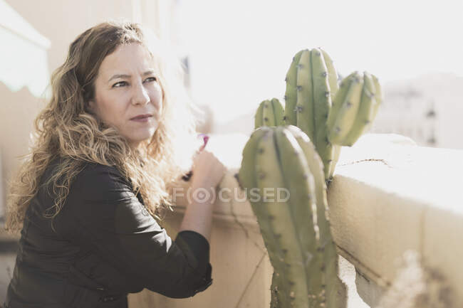Jeune dame élégante et joyeuse en robe avec la main sur la hanche près de cactus et posant sur le balcon dans la journée ensoleillée — Photo de stock