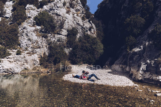Vista lateral do jovem casal deitado na costa rochosa do rio da montanha perto do penhasco — Fotografia de Stock