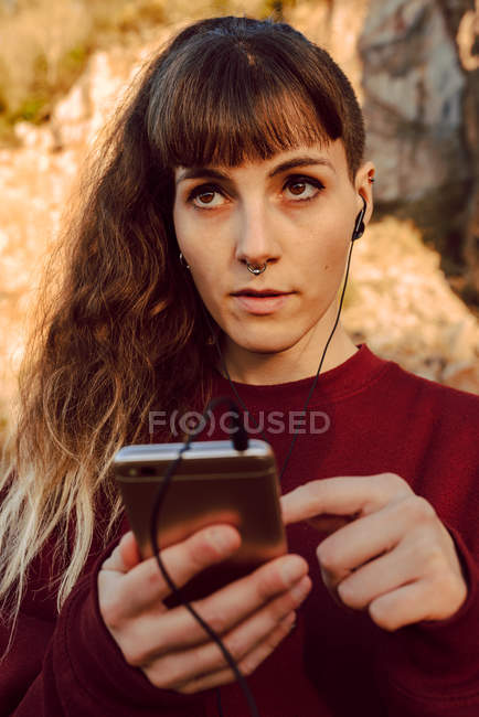 Junge Hipster-Frau mit Piercing und Kopfhörer, die auf dem Land mit dem Handy Musik hört — Stockfoto