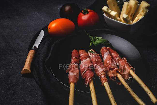 Gressini con prosciutto tipico spagnolo serrano su piatto nero con pomodori freschi e formaggio — Foto stock