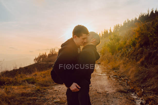 Feliz pareja homosexual abrazando y besándose en ruta en el bosque en día soleado - foto de stock