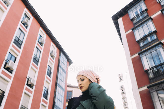 Снизу уверенная женщина с макияжем и покрытой головой позирует на улице против городских красных зданий — стоковое фото