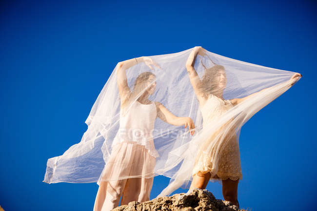 Dal basso giovani donne misteriose con le mani alzate in possesso di tessuti bianchi e in posa su rocce e cielo blu — Foto stock