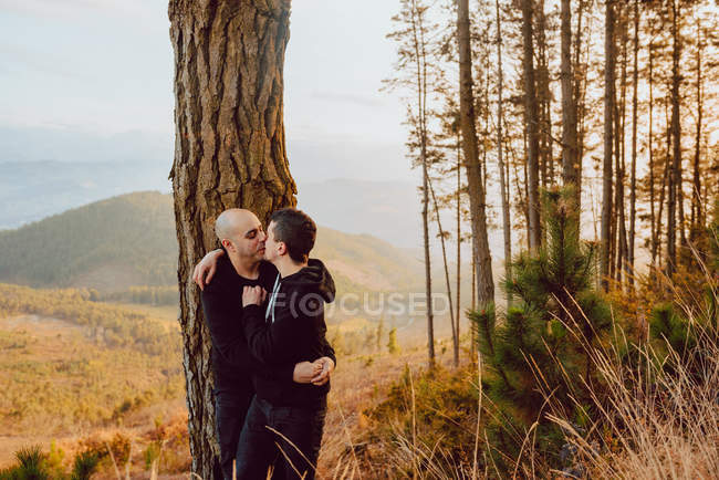 Cheerful homosexual couple kissing near tree in forest and picturesque view of valley — Stock Photo