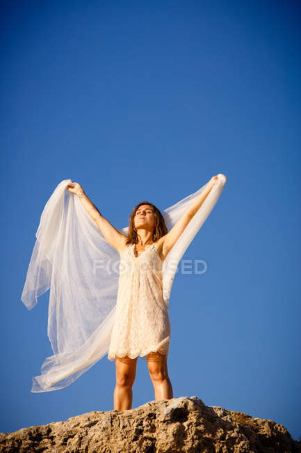 Desde abajo joven misteriosa mujer con las manos levantadas sosteniendo textil blanco y posando sobre rocas y cielo azul - foto de stock