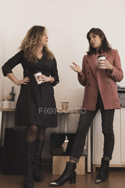 Side view of attractive young cheerful colleagues having conversation and holding mugs of beverage near printer in office — Stock Photo
