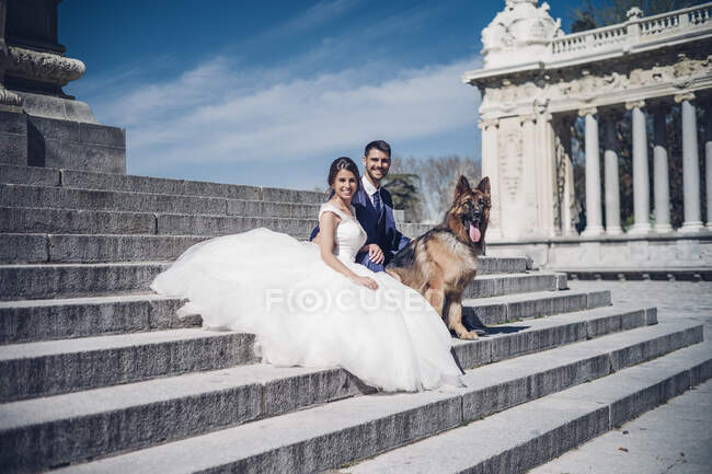 Joven pareja alegre elegante en vestidos de novia cerca de perro sentado en las escaleras cerca de edificio antiguo con columnas en día soleado - foto de stock
