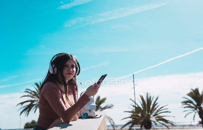 Vista lateral de mujer en auriculares con smartphone apoyado en pasamanos y mirando hacia otro lado en el ocio - foto de stock
