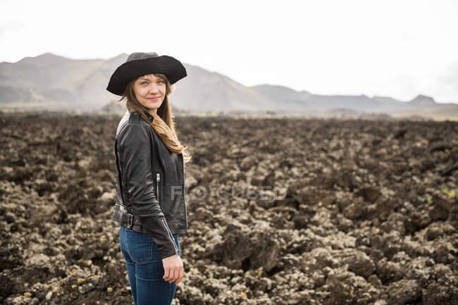Side view of attractive female in casual outfit and hat smiling and looking at camera while standing on rocky ground in majestic countryside — Stock Photo