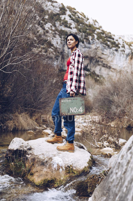 Вид сбоку случайной женщины, несущей чемодан и прыгающей по камням чистой воды в природе — стоковое фото
