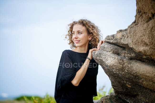 Молодая улыбающаяся женщина, опирающаяся на скалу в природе и отворачивающаяся — стоковое фото