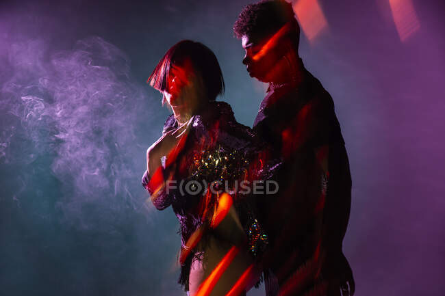 Vista lateral del atractivo hombre y mujer bailando sensual danza cerca del humo bajo brillante iluminación - foto de stock