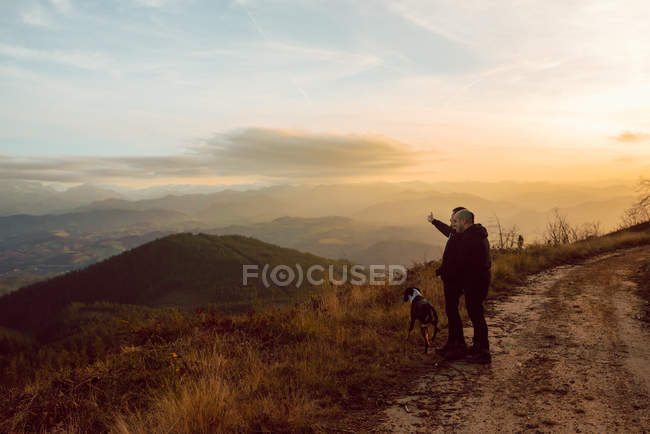 Casal homossexual de pé com o cão na rota em montanhas ao pôr do sol — Fotografia de Stock