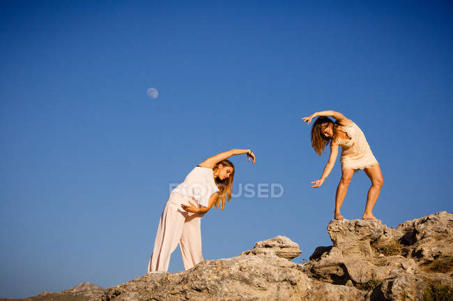 Jeunes femmes mystérieuses aux mains levées posant sur des rochers près d'une colline et ciel bleu avec lune — Photo de stock