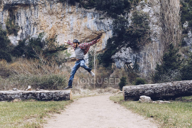 Casual man jumping among fallen trunks on rural road against beautiful cliff — Stock Photo