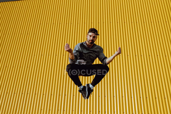 Guapo barbudo en traje de moda saltando contra la pared amarilla del edificio moderno - foto de stock