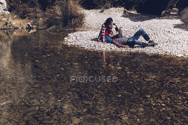 Вид сбоку на молодую пару, лежащую на скалистом берегу горной реки возле скалы — стоковое фото