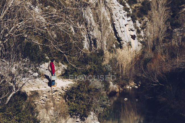 Женщина с рюкзаком и футляром стоит на скалистом берегу озера в величественных диких скалах — стоковое фото