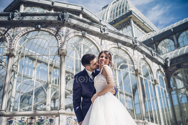 Desde abajo joven hombre elegante abrazar a la mujer en vestido de novia cerca de palacio retro con muchas ventanas en el día soleado - foto de stock