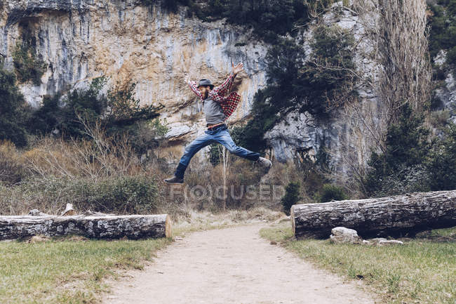 Casual man jumping among fallen trunks on rural road against beautiful cliff — Stock Photo