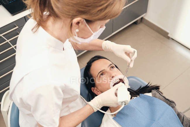 Mujer en guantes y máscara utilizando equipos modernos para la exploración de los dientes de la paciente femenina en el consultorio del dentista - foto de stock