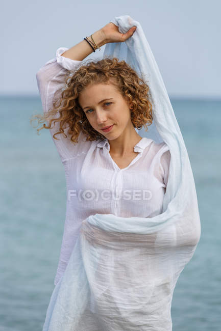 Jovem mulher olhando para a câmera e segurando lenço na mão levantada contra a superfície da água do mar — Fotografia de Stock