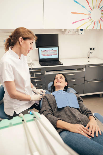 Dentista professionista in uniforme che parla con una donna sorridente mentre lavora in una clinica moderna — Foto stock