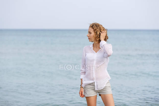Joven mujer encantadora en camisa blanca de pie en agua de mar y mirando hacia otro lado - foto de stock