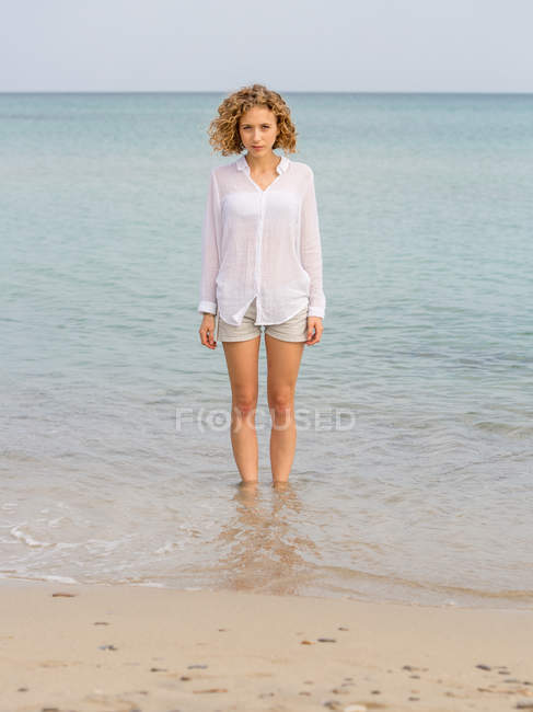 Joven mujer encantadora en camisa blanca caminando en agua de mar y mirando a la cámara - foto de stock