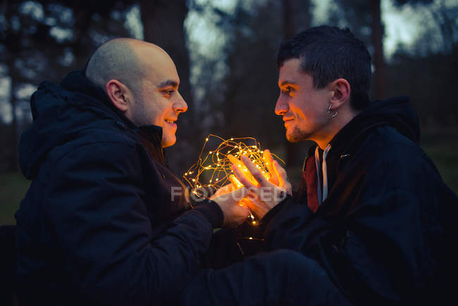 Pareja homosexual con luces de hadas iluminadas sentada en el bosque oscuro por la noche - foto de stock
