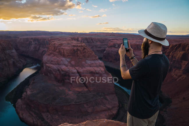 Handsome man in hat taking picture while standing against magnificent canyon and river during sunset on West Coast of USA — Stock Photo
