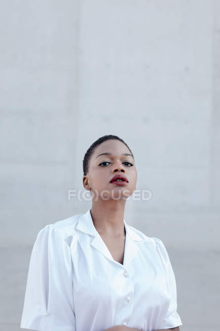 Moda modello donna etnica dai capelli corti in camicia bianca in posa contro parete grigia — Foto stock