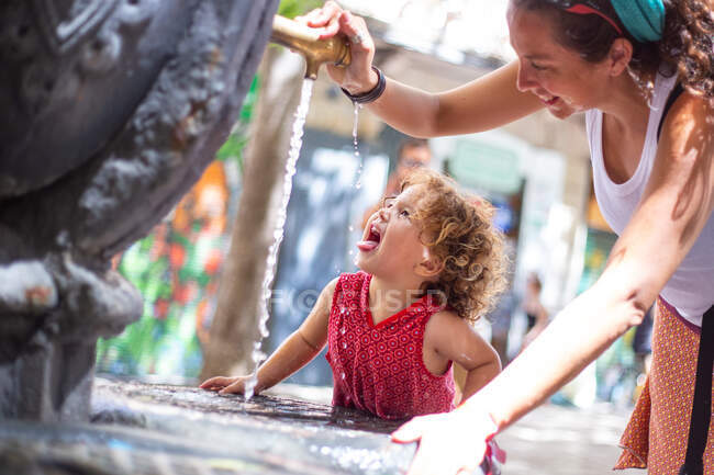 Felice madre versando acqua da bere fontana per divertente ragazza con bocca aperta sulla strada in estate — Foto stock