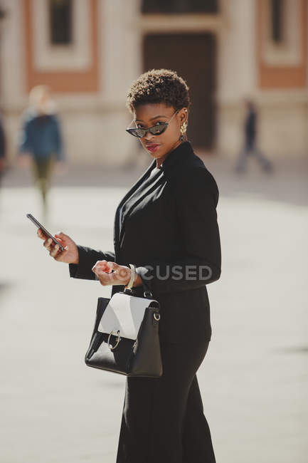 Confiante afro-americano elegante mulher em traje e óculos de sol segurando saco e telefone celular na rua — Fotografia de Stock