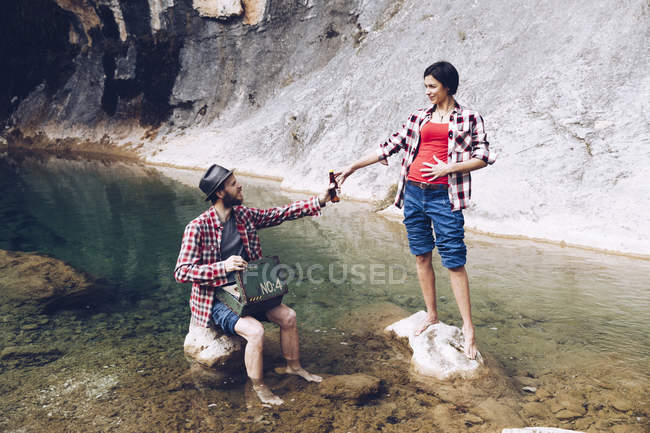 Чоловік і жінка у прозорій воді красивого озера на каменях з пляшкою пива насолоджуються пікніком — стокове фото