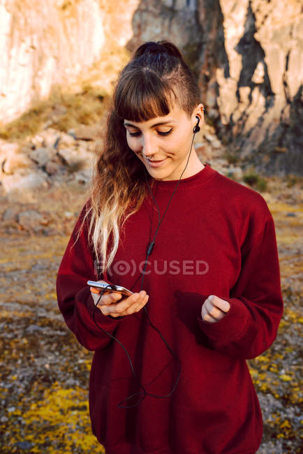 Jeune femme hipster avec perçage et écouteurs écoutant de la musique avec téléphone portable et marchant sur la route de campagne — Photo de stock