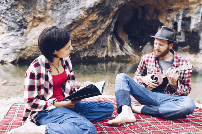 Homme et femme adultes sur carreaux à carreaux avec livre et petit ukulélé pique-nique sur la rive du lac — Photo de stock