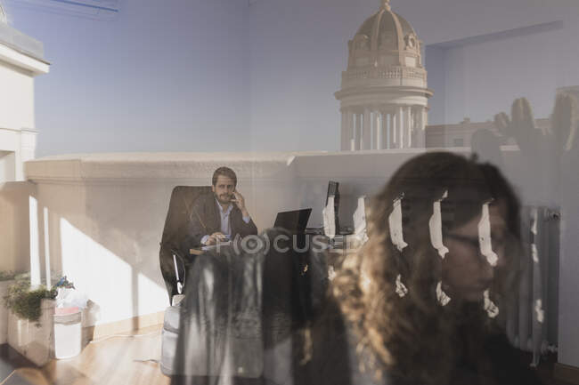 Concentrated young female and male talking on mobile phone and browsing on laptop at tables in office through window — Stock Photo