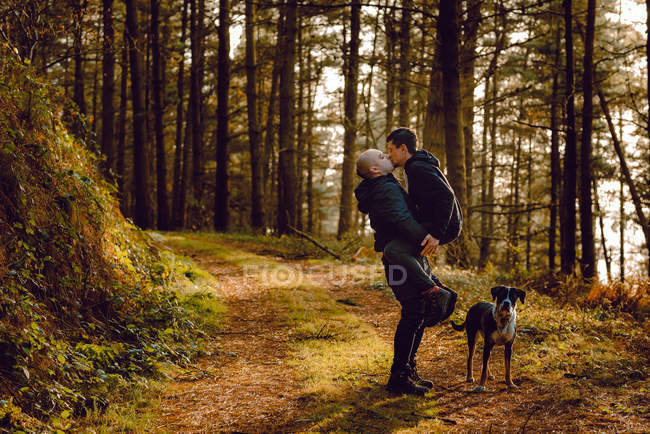 Casal homossexual abraçando perto do cão na rota na floresta em dia ensolarado — Fotografia de Stock