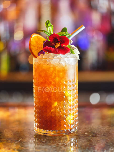 Glas leckeren Cocktails mit roten Blüten und Scheiben frischer Zitrusfrüchte dekoriert und auf Theke auf verschwommenem Hintergrund der Bar platziert — Stockfoto