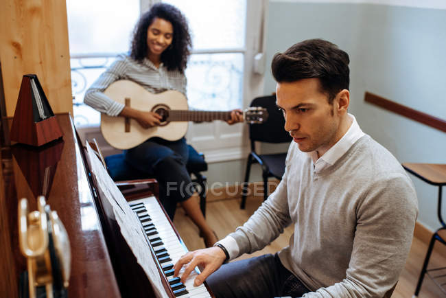 Vue latérale du jeune homme jouant du piano près de la femme noire jouant de la guitare en studio de musique — Photo de stock