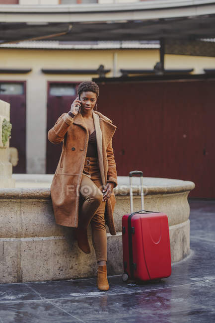 African American elegant woman with baggage talking on mobile phone and standing near monument on street — Stock Photo