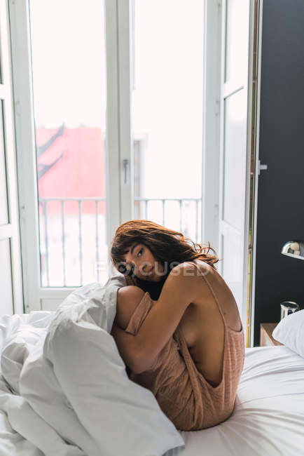 Young cozy woman in nude dress sitting on bed in bedroom — Stock Photo