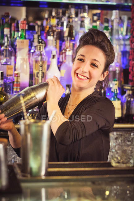 Jolie femme barman souriant et mélangeant cocktail dans shaker tout en se tenant derrière le comptoir dans le bar et en regardant loin — Photo de stock