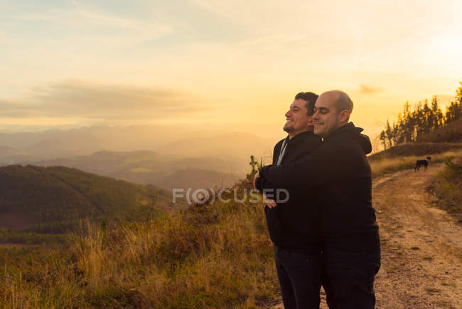 Feliz casal homossexual abraçando e desfrutando do pôr do sol na rota nas montanhas — Fotografia de Stock