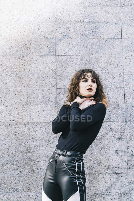 Attractive young woman in stylish outfit choking herself and looking away while standing near building wall on city street — Stock Photo