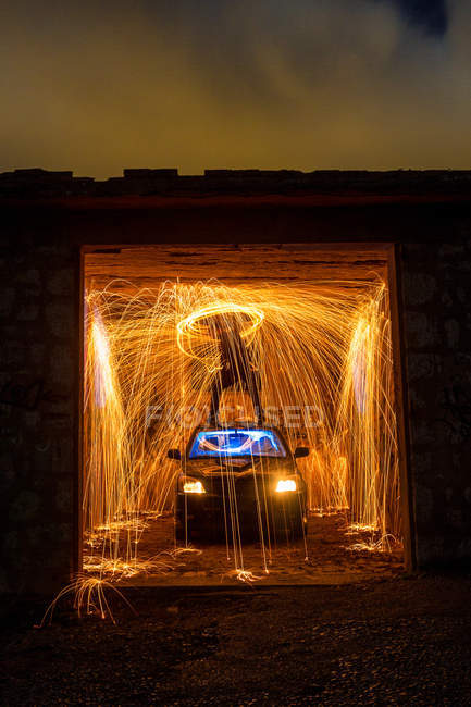 Длительное обнажение силуэта человека с горящим кругом, стоящим на крыше автомобиля в сарае между фейерверками в темноте — стоковое фото